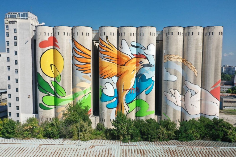 A monumental mural created on the façade of the largest grain storage facility in Europe, located in Sofia, Bulgaria. The artwork covers approximately 4,000 square meters and rises to a height of 42.5 meters, transforming an industrial complex into a powerful artistic symbol visible from a great distance.

At the center of the composition is a bird in flight, connecting sky and earth. Its outstretched wings echo waves of air and clouds, while nearby appear images of grain and human hands—symbols of labor, care, and the continuity of life. Together, these elements form a visual metaphor for the interconnection between humanity, nature, and the cycles of harvest.

The work balances the monumental scale of industrial architecture with a soft, living language of symbols. The mural speaks of growth, hope, and forward movement, transforming a space of storage into a space of meaning—where past, present, and future are united in a single gesture of flight.