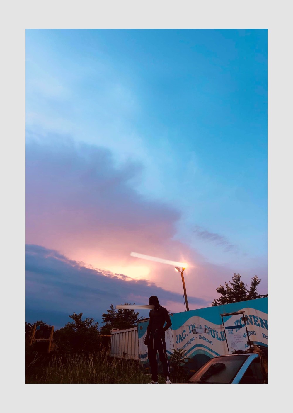 In this captivating piece titled "Rebel Against the Dusk," the composition captures a lone figure standing beneath an expansive, fading sky, where the hues of pink, purple, and blue meld into twilight. The contrast between the dark, shadowed silhouette and the ethereal, almost dreamlike colors of the sunset creates a striking tension a balance between light and darkness, freedom and solitude.
The figure’s stance, positioned in front of an industrial, almost abandoned-looking backdrop, speaks of resilience and individuality, seemingly unyielding in the face of encroaching shadows. The use of light, with the single streetlamp casting a soft glow over the scene, reinforces a sense of rebellious energy against the approaching night, adding a layer of mystery.
This artwork embodies themes of solitude, resilience, and defiance against the constraints of both time and place.

Created By Kwaku Kr3