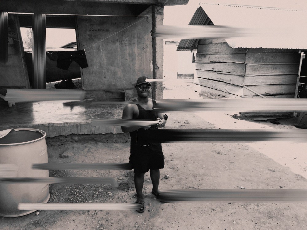 This evocative artwork explores the concept of fragmented identity and self-perception in a rapidly changing world. The image, presented in a muted monochromatic tone, juxtaposes the solidity of a rural setting with the distortion of the subject’s figure through dynamic streaks of motion. The man at the center, grounded yet partially obscured, represents the human experience of being caught between different dimensions of self-awareness, memory, and societal roles.

The deliberate blurring and streaking effect symbolize the layers of consciousness that often overlap, conceal, or redefine our identity. Each streak could be seen as a representation of past experiences, fleeting emotions, or external influences, challenging the viewer to question what remains constant amid life’s chaos.

The rustic surroundings a house under construction, weathered wooden structures, and a barrel add to the narrative of transformation and resilience. The environment mirrors the subject’s own journey.