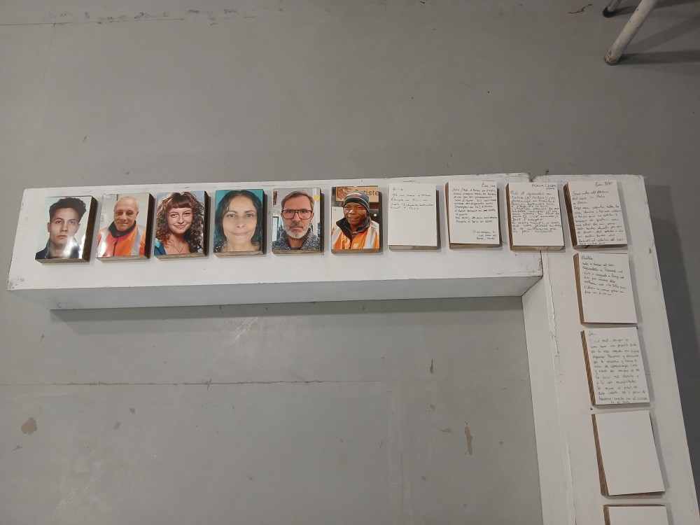 Détail de l'installation à Shakirail à Paris, lors de l'exposition RAMIFICATIONS, novembre 2025. Portraits photographiques couleur sur papier glacé 10x13, contrecollés sur un support en bois. Fiches blanches portant des témoignages écrits de visiteurs relatant leur expérience de migrants, rédigés lors du vernissage. L'œuvre se poursuit par la collecte d'images, de vidéos, d'entretiens et de textes.