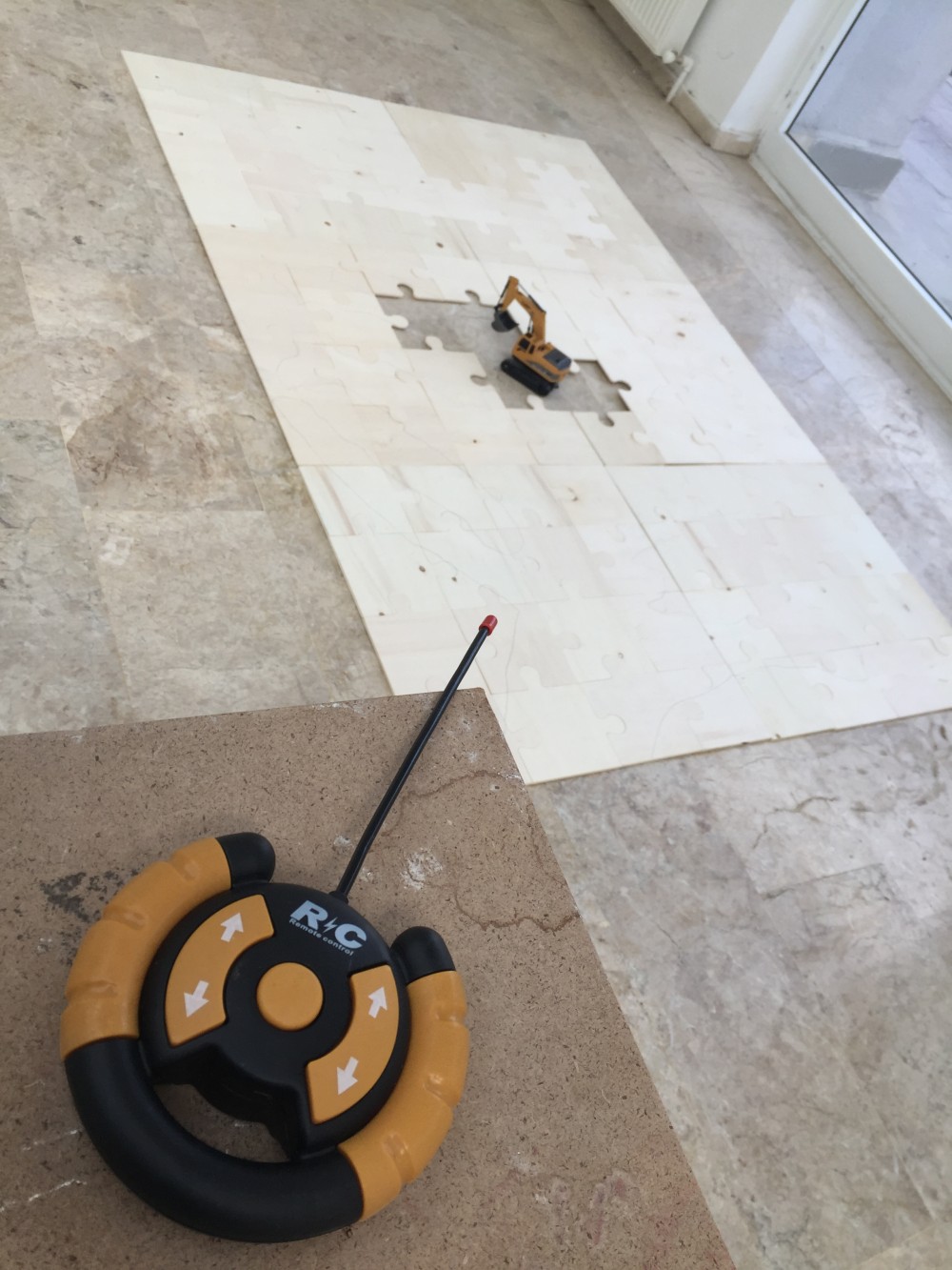 This installation addresses the urban transformation of Dikmen Valley (4th and 5th stages) in Ankara, where informal settlements were demolished and replaced by profit-driven high-rise developments. These changes displaced low-income residents, erased public and ecological spaces, and intensified income inequality.

Six wooden puzzles, each composed of 16 pieces, are arranged to form a rectangle. Several central pieces are removed, creating a void that contains a remote-controlled excavator. A pencil-drawn site sketch of Dikmen Valley is rendered across the puzzle surface.

The excavator acts as a destructive agent controlled remotely, symbolizing power and authority, while each puzzle piece represents a parcel of land subject to speculation. Trapped within the void, the excavator repeatedly strikes the surface, emphasizing the aggressive and cyclical nature of profit-driven urban transformation.