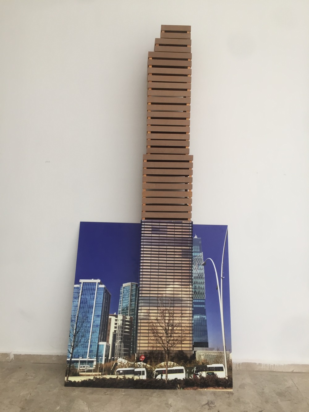 Kasa Plaza narrates the condition of individuals who are subjected to an imposed way of life and stacked within isolated spaces, where uniformity becomes a mode of survival. Within these multi-storey structures—devoid of spaces for encounter—class distinctions among urban residents become increasingly apparent. The crates, objects of consumer culture whose visual form recalls skyscrapers, are transformed into endlessly rising architectural floors within the work.

The installation is composed of three distinct parts and is given a three-dimensional spatial effect. Archival images of skyscrapers are printed onto composite plates positioned in front of, to the right, and to the left of the crates, forming a spatial layer that merges with them. In this configuration, the skyscraper becomes both the base and an integral component of the crates, collapsing architectural representation and lived space into a single structure.